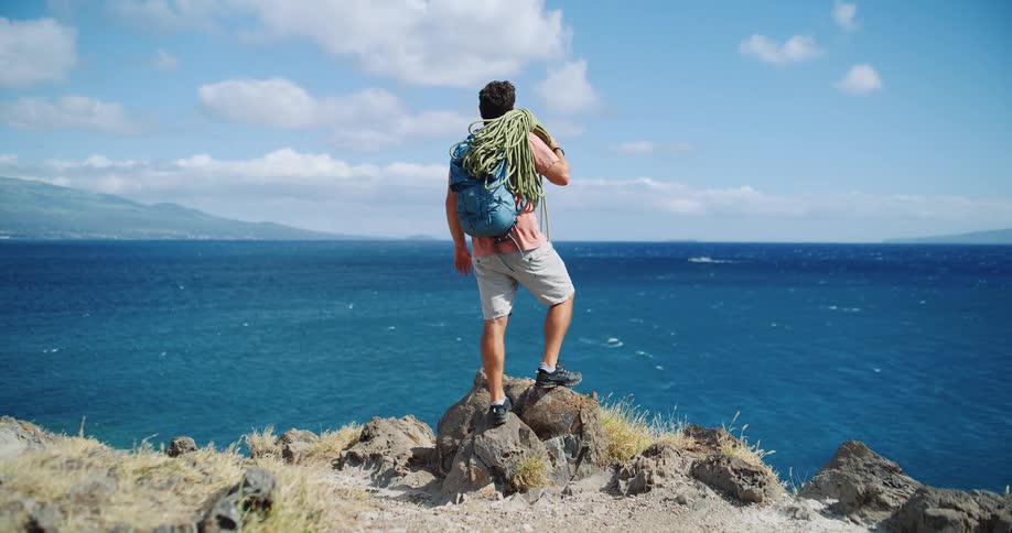 A man stands on a rocky cliff overlooking the ocean with hiking gear