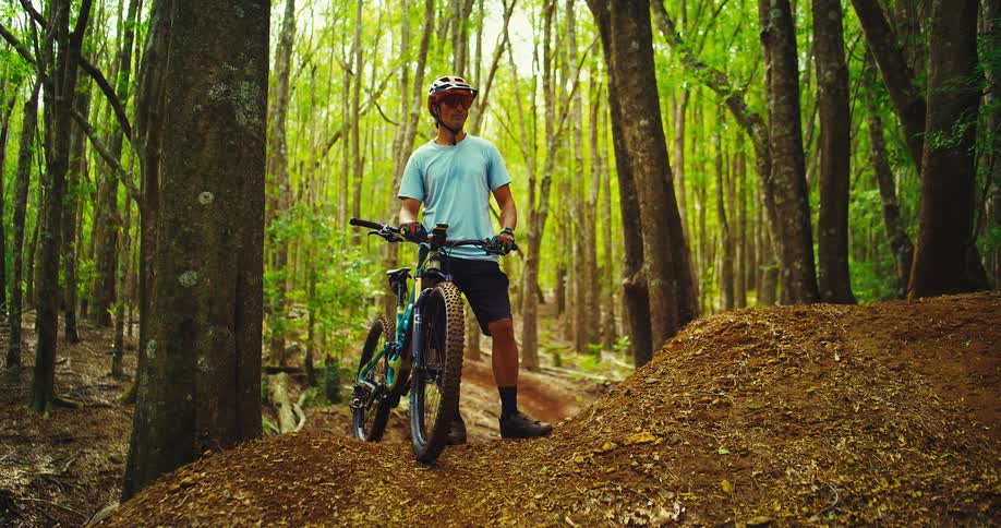 A mountain biker is standing on a dirt trail in the middle of a forest he is wearing a helmet and a light blue shirt holding onto the handlebars of his bike