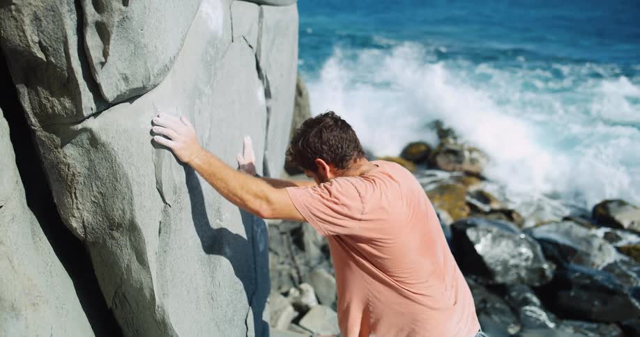 Man climbing a seaside rock with waves crashing against the stones in the background