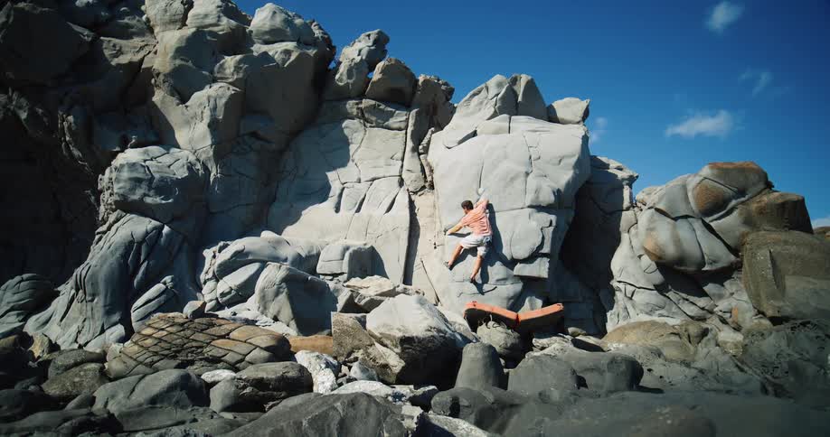 The video shows a person rock climbing on a large rugged rock face the climber is wearing an orange shirt and shorts and an orange crash pad is visible below them