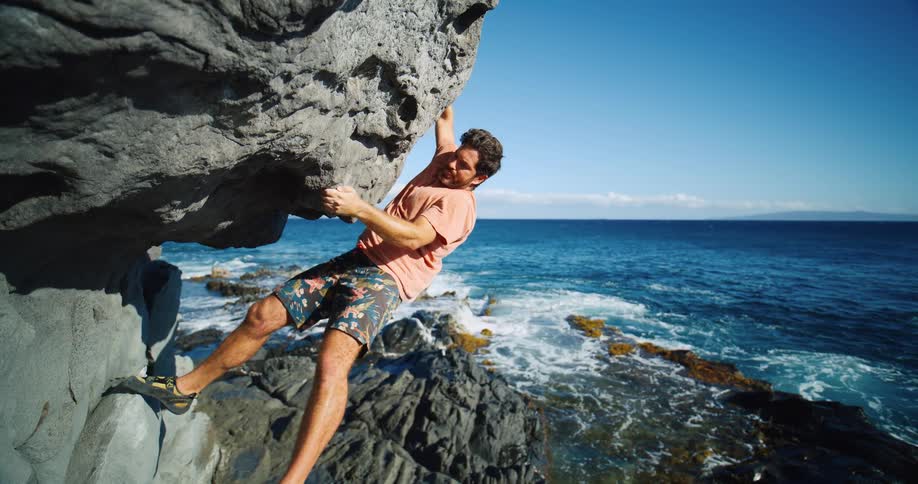 A man is seen rock climbing on the side of a cliff face the ocean and a small island are visible in the background
