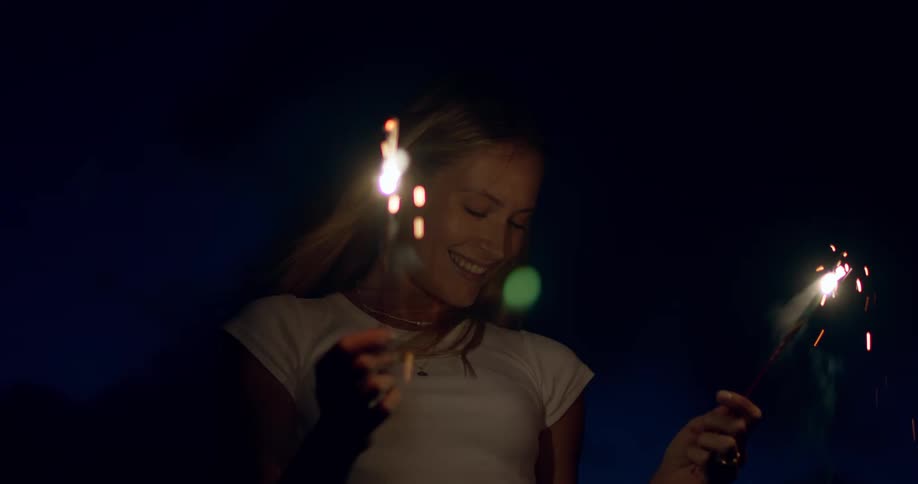 A blonde woman stands in the dark holding two lit sparklers shes wearing a white tshirt and smiling