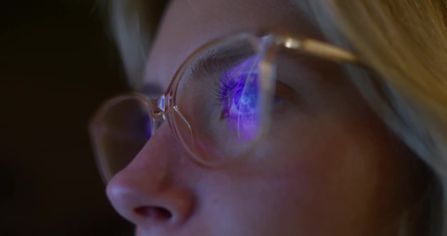 A close view shows a woman with blonde hair and a pale complexion wearing clear wire rim glasses as the bright blue light of a screen reflects on the surface of her lenses