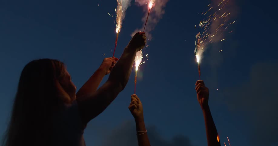 Friends raise sparklers into a deep blue evening sky sparks trailing upward