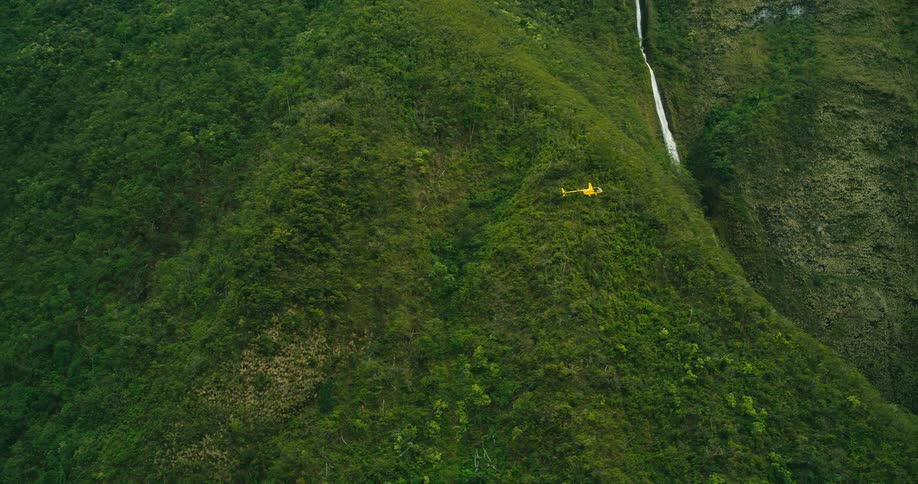 A yellow helicopter flies over a lush green mountain