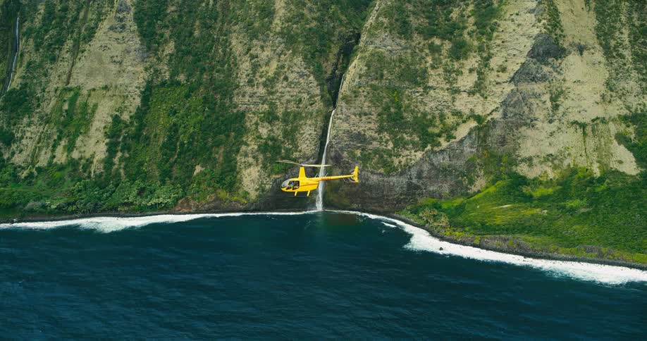 Wide aerial view of a bright yellow chopper flying along towering green cliffs where a thin waterfall drops to the blue ocean below