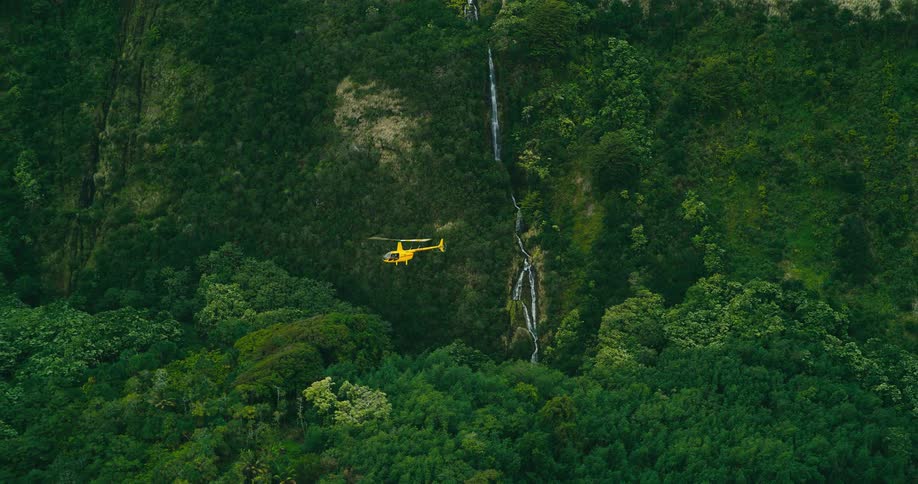An aerial tracking shot follows a bright yellow helicopter as it flies past a magnificent waterfall on a lush junglecovered mountainside