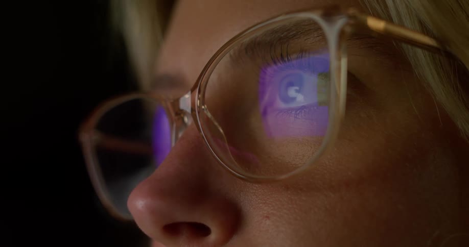 A closeup shot shows a person wearing eyeglasses in a dark room the reflection of a bright glowing square with a glowing icon in the center is visible on the lenses of the glasses