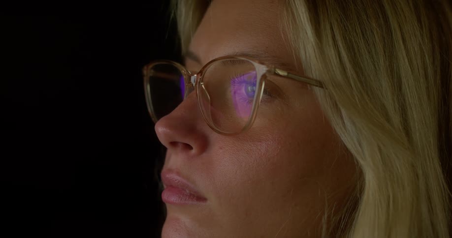 The face of a blonde woman wearing glasses that are reflecting a purple light on the lens in a dark room