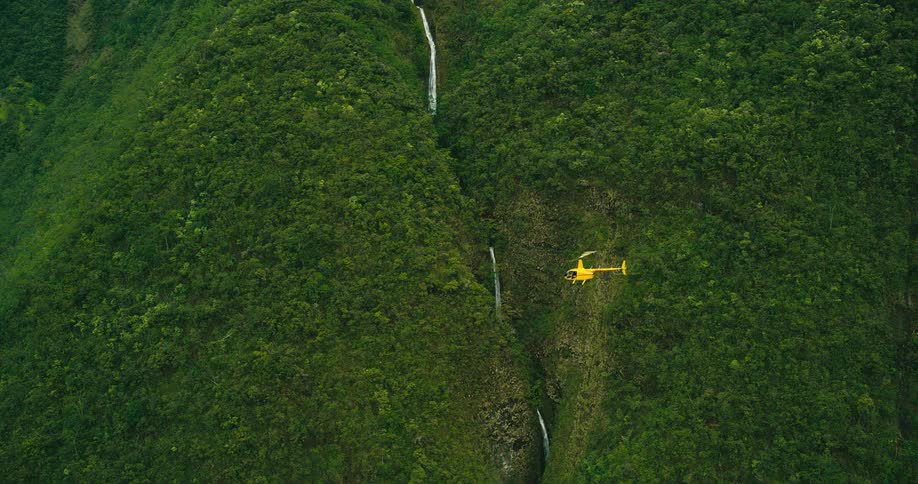 An aerial shot follows a small yellow helicopter as it flies through a vast canyon with a waterfall and lush green mountain slopes