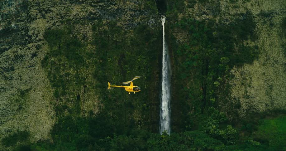 A yellow helicopter flies between two tall waterfalls along a rugged cliffside