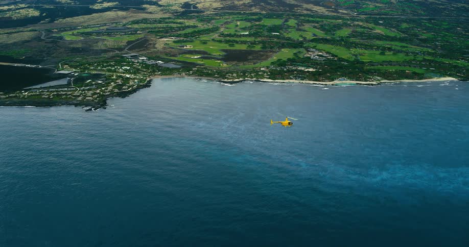 A yellow helicopter flies over a lush coastal landscape beside the ocean