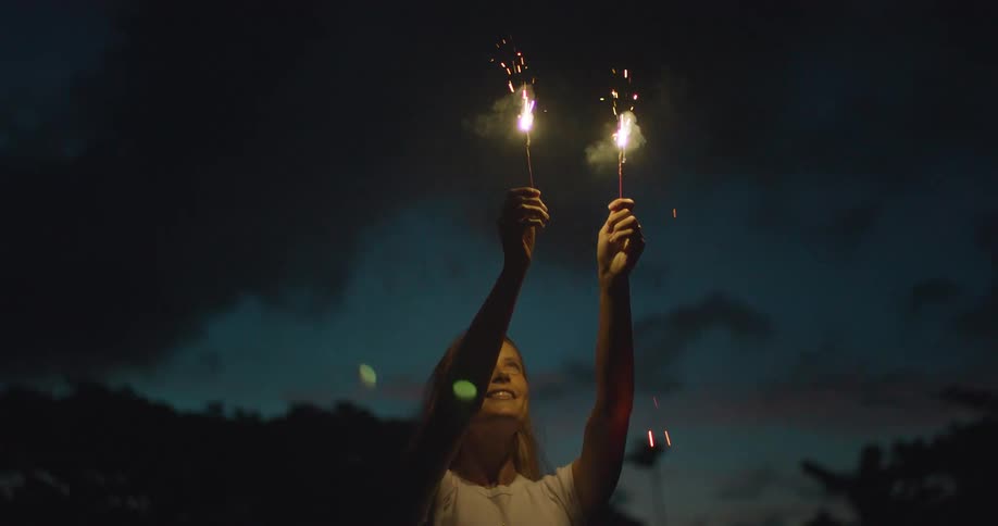 A woman holding two lit sparklers outdoors at night
