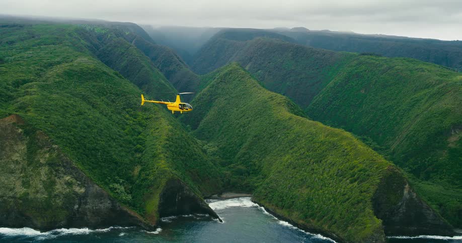The video shows a bright yellow helicopter flying over a lush green valley with steep slopes leading down to the ocean the helicopter hovers and then flies forward showcasing the dramatic mountainous landscape