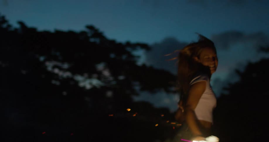 A young woman dances and runs outdoors while holding fireworks in both hands at night