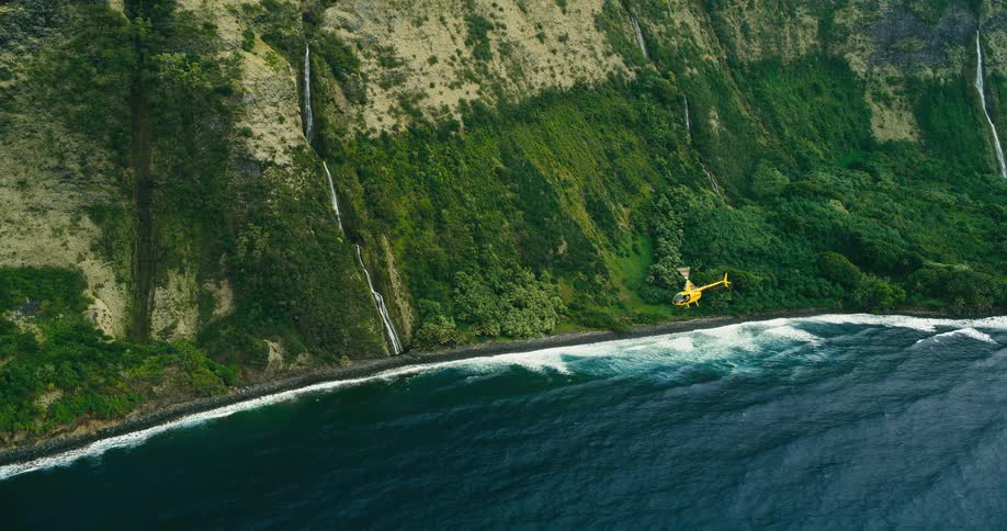 A yellow helicopter is flying over the water of a mountainous coast