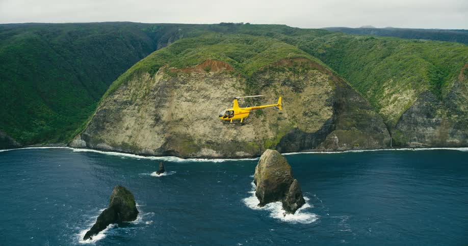 A yellow helicopter flies on a scenic tour past massive greentopped sea cliffs and rock formations rising from the deep blue ocean