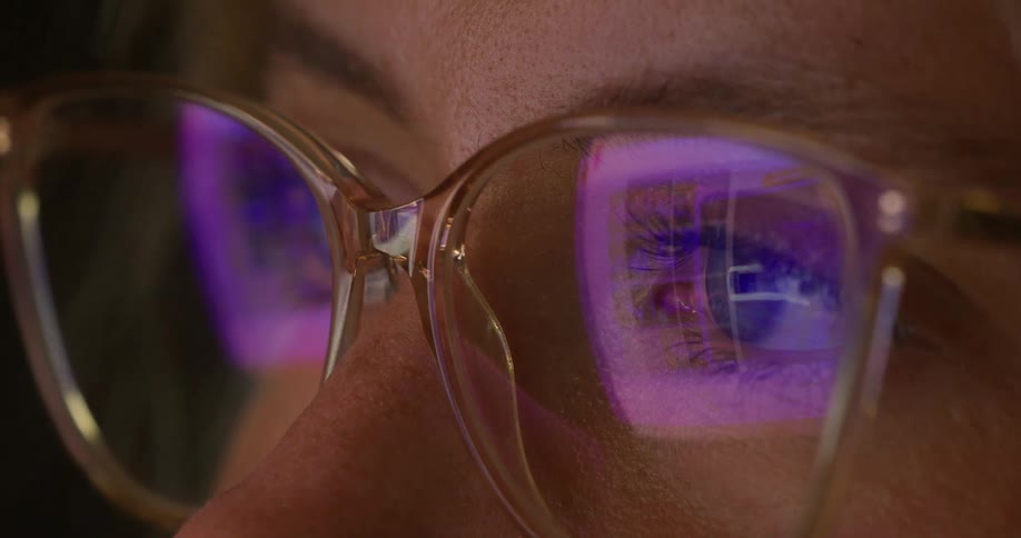 The face of a woman wearing glasses that are reflecting a purple light and she is looking straight ahead blinking her eyes