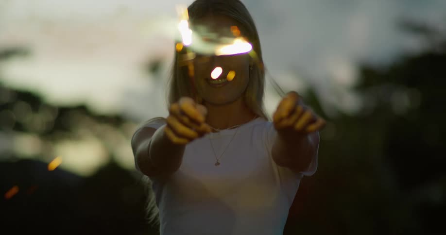 A closeup shot of a happy woman with blonde hair smiling as she holds and playfully moves two lit sparklers in front of her