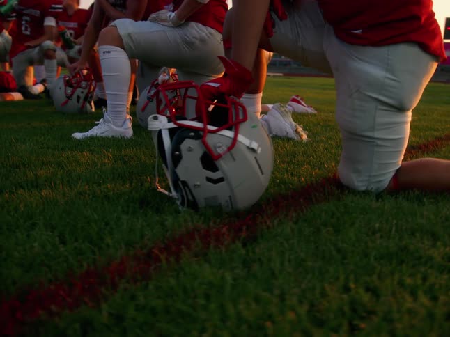 American football players kneel on a grassy field at sunset with their helmets placed on the ground