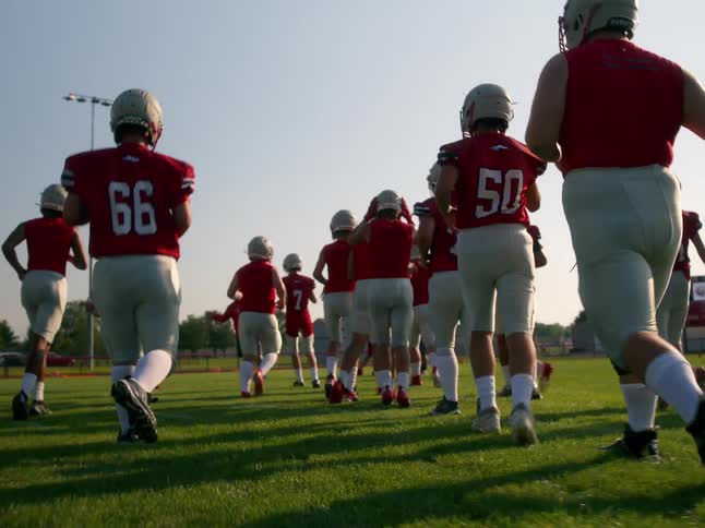 Football players run onto the field at midday wearing red jerseys and helmets