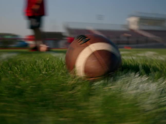 A football rests on green turf as a blurred player stands in the background