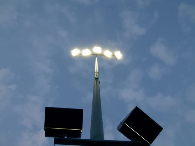 A tall stadium floodlight shines brightly against a blue sky