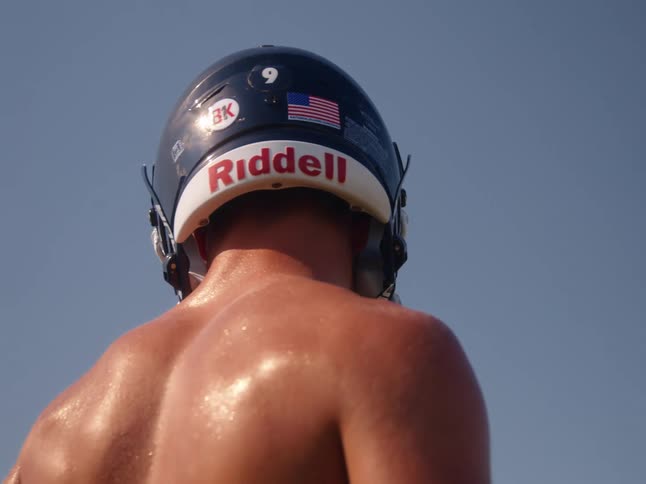 A shirtless football player wearing a helmet is seen from behind standing on a field the camera then zooms in on the back of his helmet which has the number 9 and an american flag sticker on it