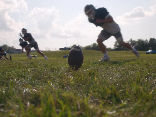 Lowangle closeup of an upright football on the field as players sprint and one player kicks the ball