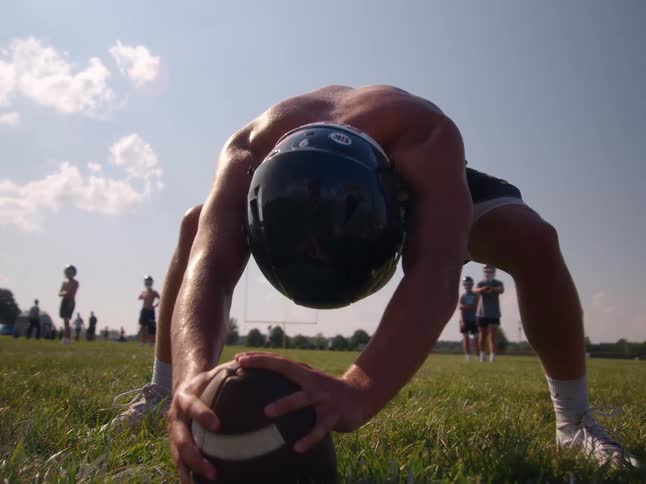 An american football player in a helmet and shorts gets ready to snap the ball on a grassy field with other players in the background
