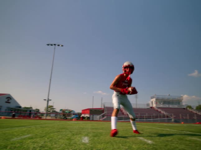 The video shows an athlete in a red jersey and white pants running on a football field he catches a football and runs toward the camera
