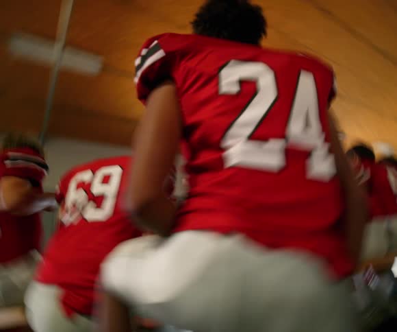 A lowangle motionblurred shot shows several football players in a locker room the players are dressed in red jerseys and white pants and one player wearing jersey number 24 stands up from a bench