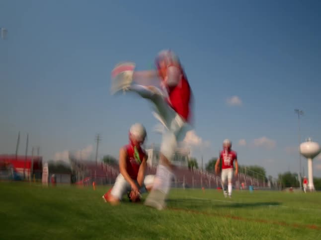 The video shows two football players in red jerseys and white pants in the first half one player is kneeling while another player runs quickly across the field in the second half of the clip a player wearing a helmet is walking towards the camera