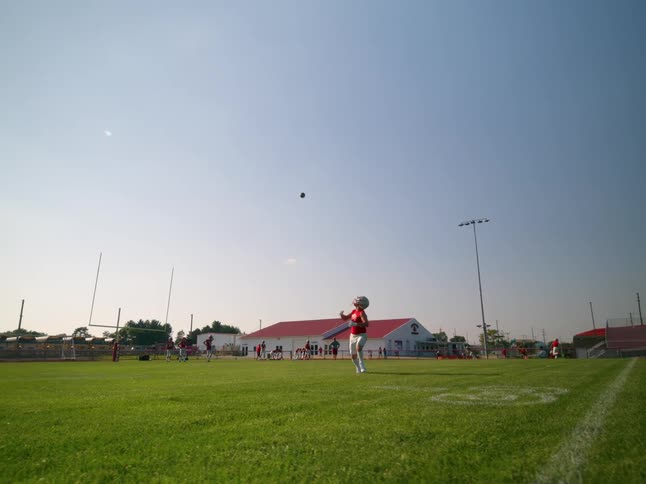A young man wearing a football helmet and a red jersey is shown running across a green football field he is looking at a football flying through the air preparing to catch it