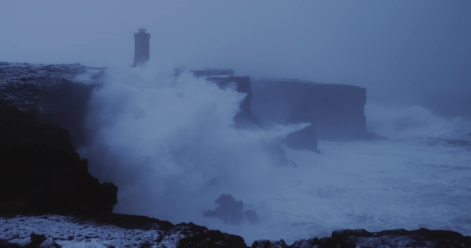 A massive wave crashes against coastal cliffs near a lighthouse