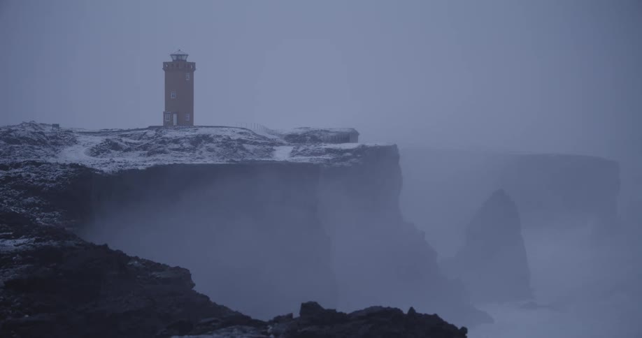 A solitary lighthouse stands on a snowy cliff above a foggy sea