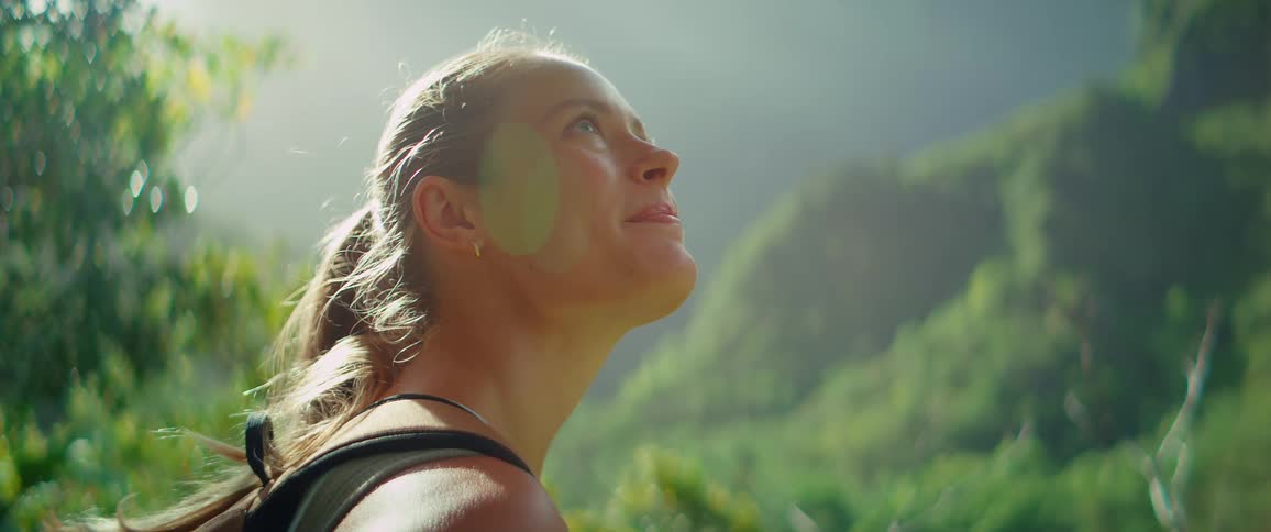 A closeup shot of a young woman with a backpack her face lit by sunlight as she looks around with a smile while hiking in a lush green mountainous area