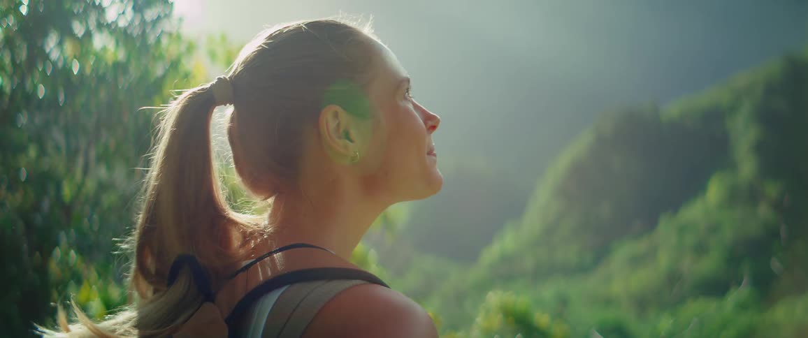A woman with her long blonde hair in a ponytail stands on a trail looking off to the right with a relaxed expression as light shines behind her and illuminates the foggy mountainous forest in the background