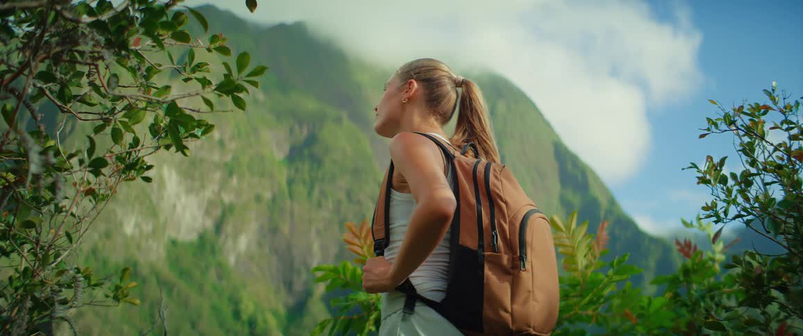A young woman carrying a backpack while hiking in the woods stands observing the forest view with a mountain in the distance at midday