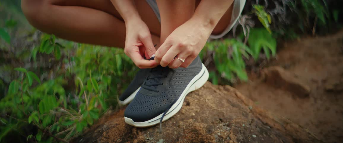 A woman wearing black running shoes with white soles crouches and ties her shoelaces on a large brown rock