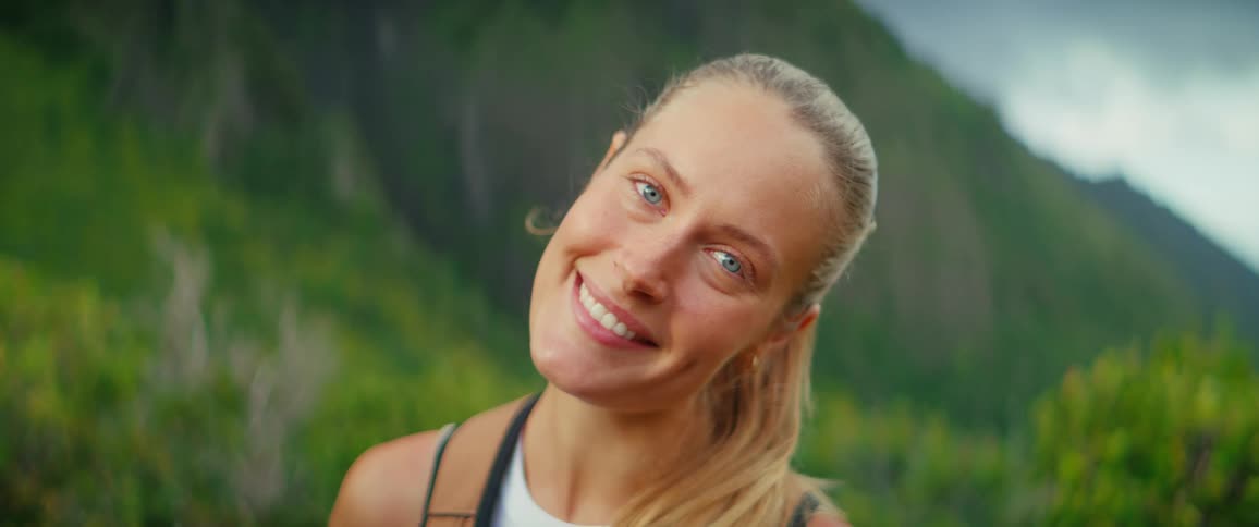 A blonde woman with her hair tied back is seen hiking she closes her eyes and then smiles at the camera showing her dimples