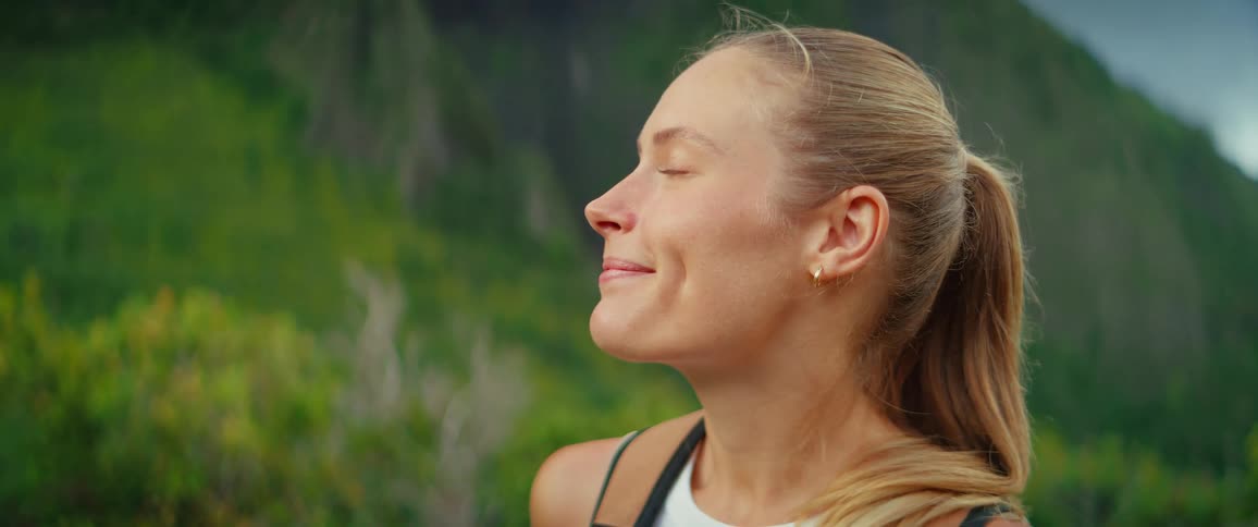 A smiling blonde woman stands outside on a hillside wearing a backpack and looking toward the left side of the frame her eyes close briefly as she appears to be enjoying the scenery and fresh air