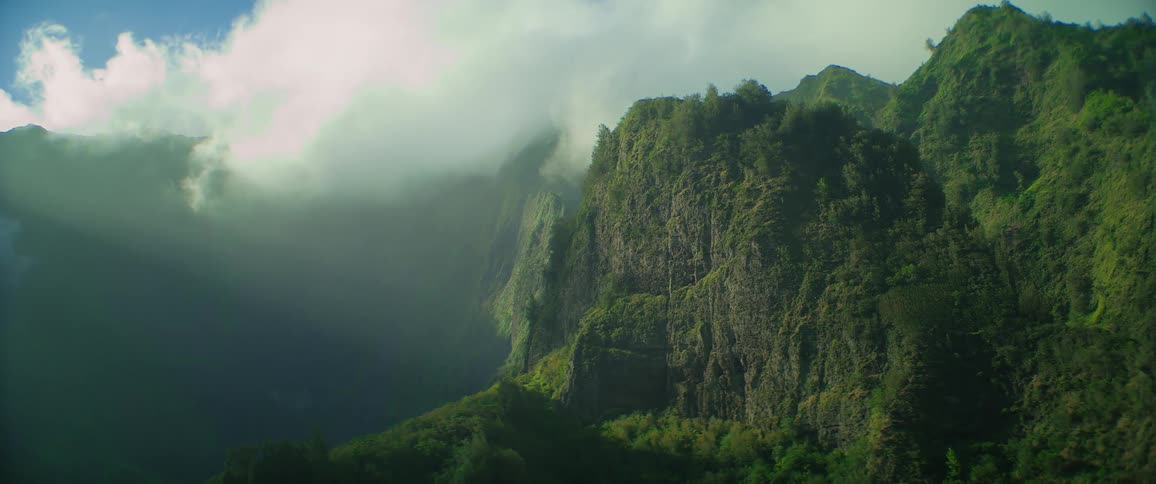 Sunlight illuminates a lush green mountain cliff through the clouds