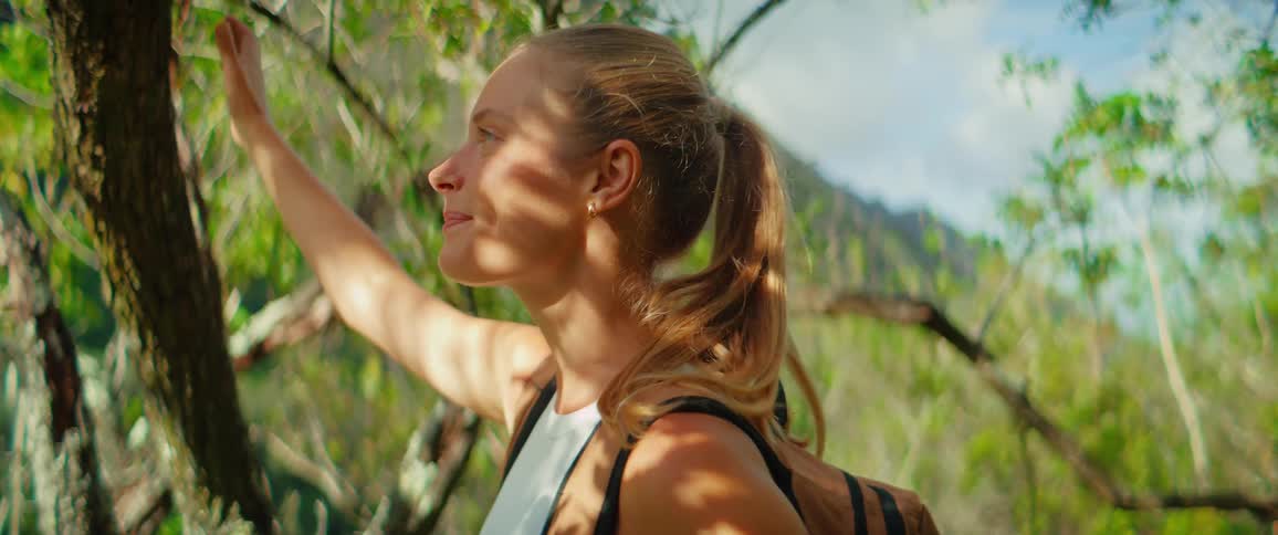 A blonde woman wearing a backpack is smiling as she hikes in a lush green forest she stops to admire the view and then leans against a tree trunk appearing to be at peace with her surroundings