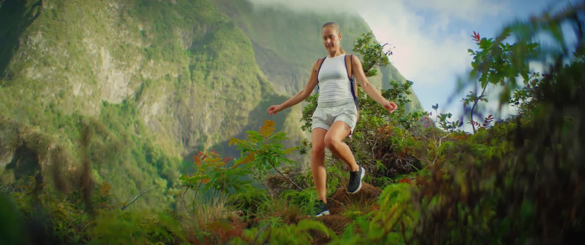 A wide shot of a woman with a backpack walking on a rocky plantcovered trail with a view of a massive green mountain range