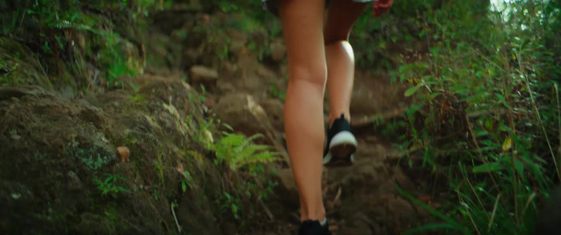 A young woman walking up a rocky path in the woods during a hike wearing shorts and carrying a backpack at midday