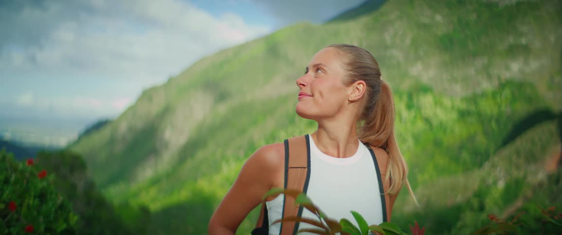 A blonde woman with her hair in a ponytail is standing on a green hillside with a backpack on she turns her head and smiles widely at the camera while wearing a white tank top and lightcolored shorts