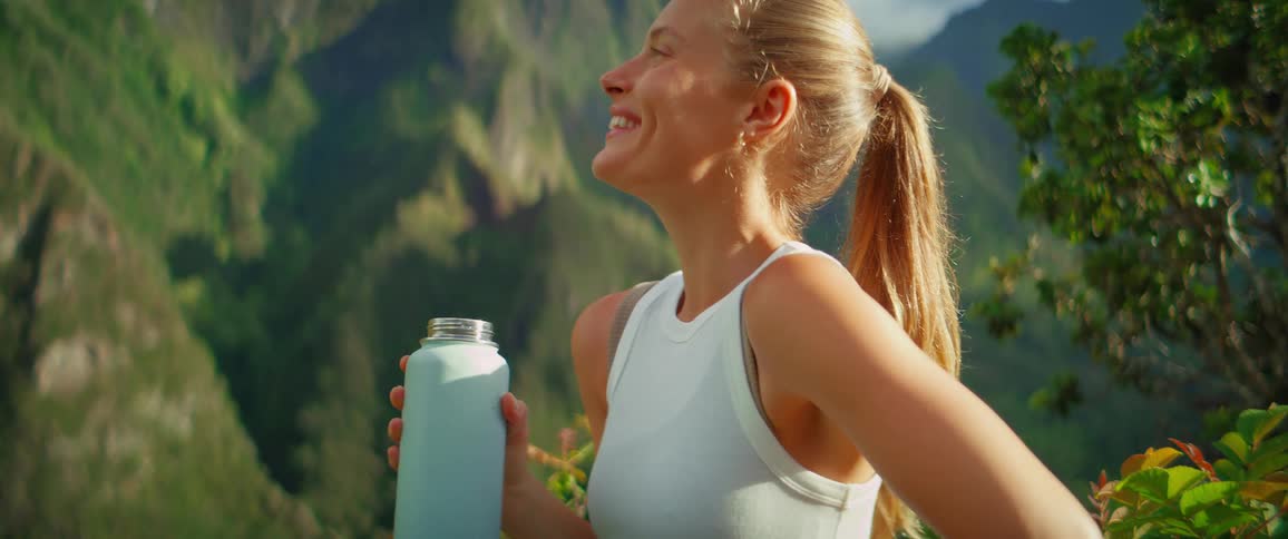 A blonde woman in a ponytail stands outdoors with a beautiful green mountain range in the background she holds a light blue water bottle and takes a long drink from it then smiles