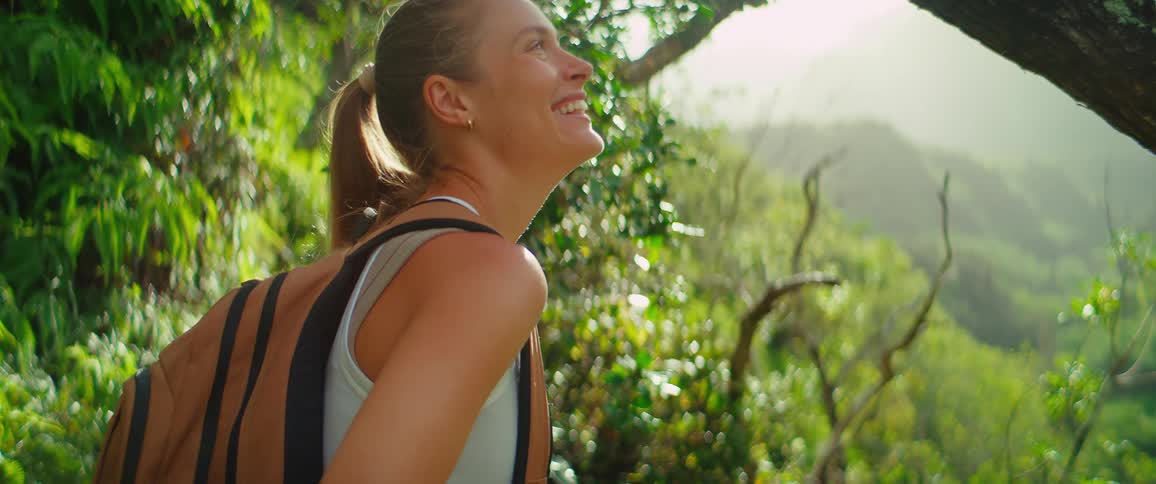 A young woman carrying a backpack standing in the woods during a hike at midday smiling