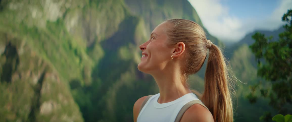 The video shows a closeup shot of a blonde woman with a ponytail holding a water bottle she is standing outdoors with lush green mountains in the blurred background she smiles looks down and then looks up and off into the distance appearing refres